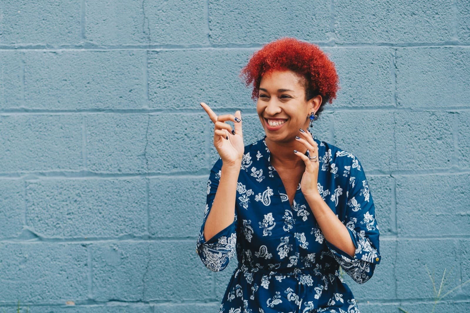 A smiling adult with bright red hair against a blue brick wall