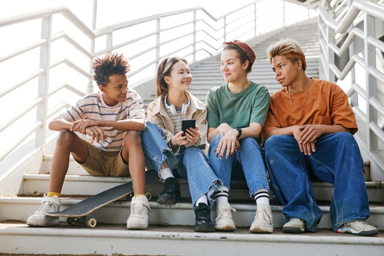 Four teens sitting together on outdoor steps, talking, one holding a phone and a skateboard nearby