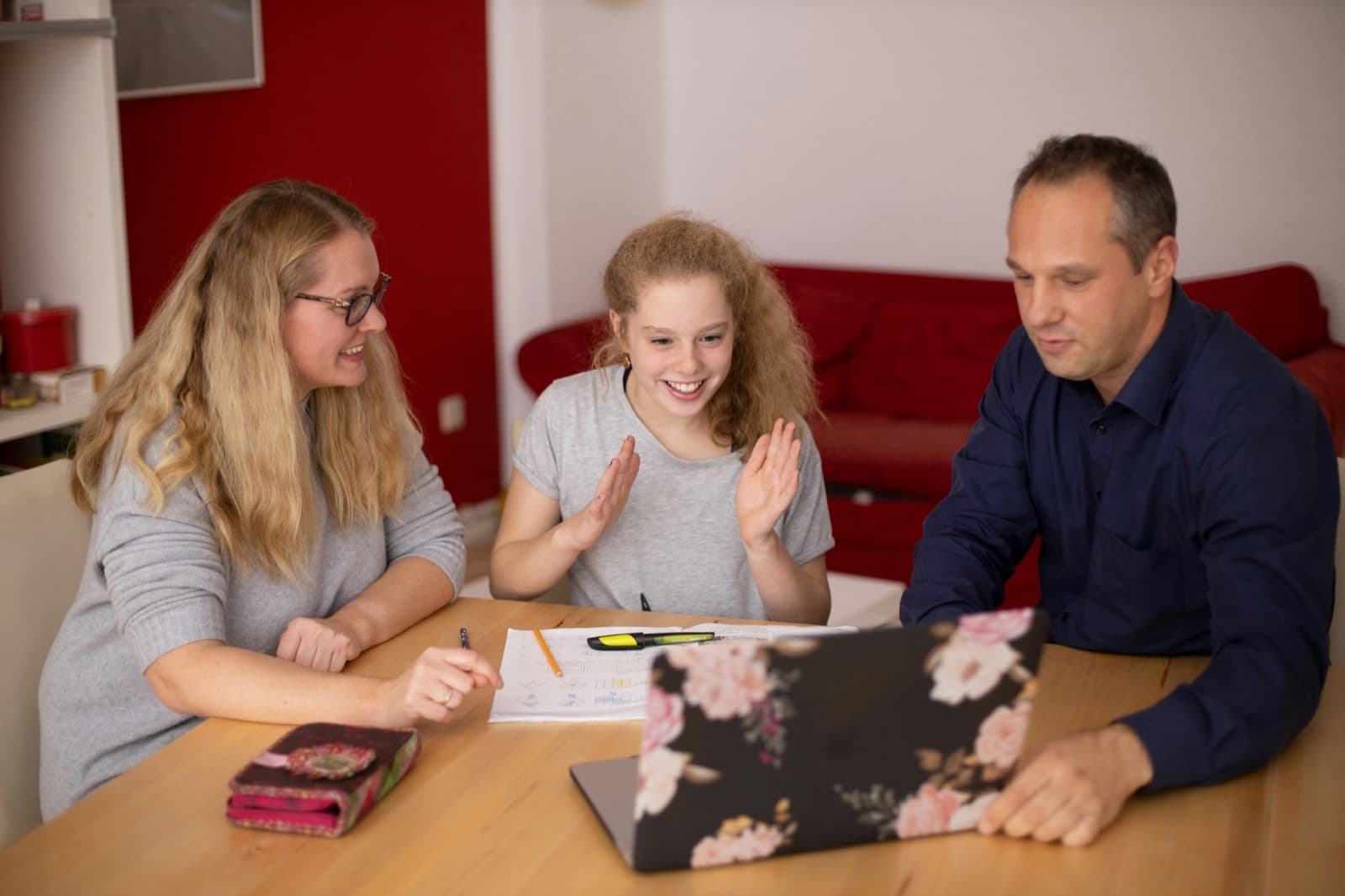 A parent and teen work together at a laptop at the kitchen table while the teen waves at the screen