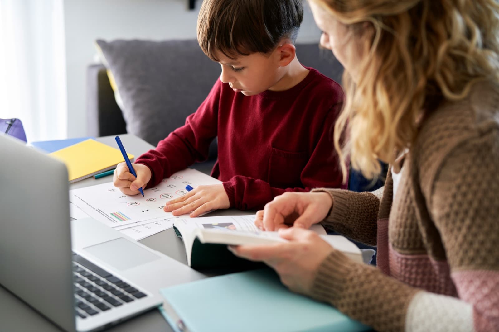 A parent reviews paperwork beside a teen writing at a desk with a laptop