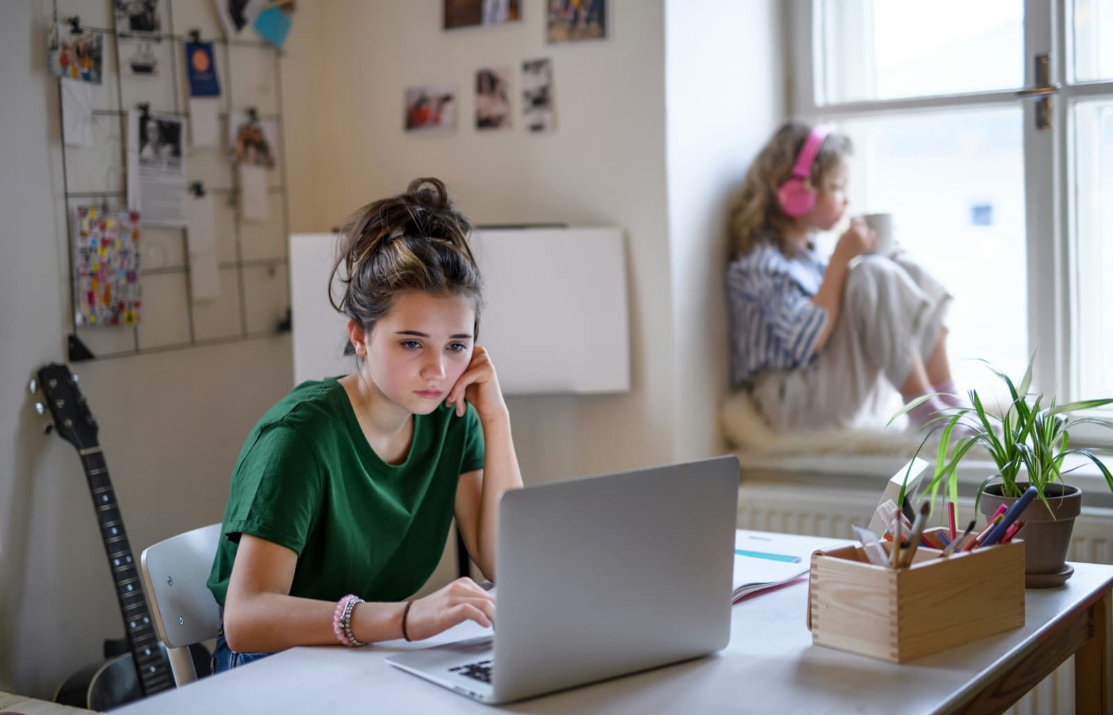 A teen working at a laptop at a desk in her bedroom while another relaxes on the windowsill behind her