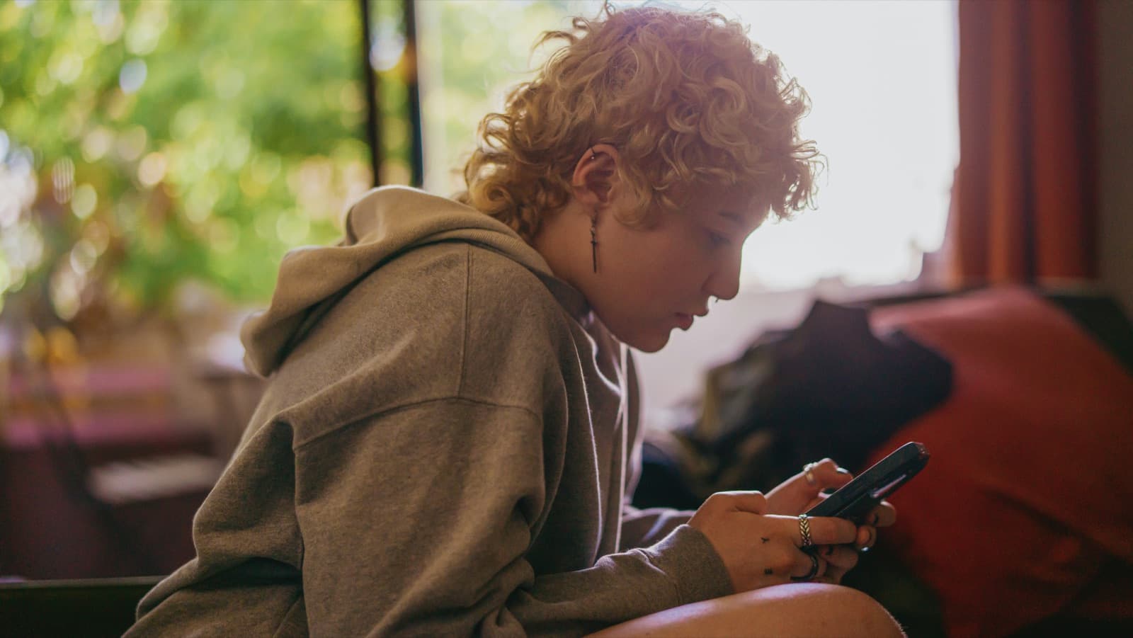 A teen with curly hair sits quietly on a couch looking at their phone